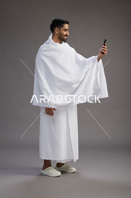 بورتريه لرجل سعودي بملابس الإحرام يبتسم ويلتقط صورة سيلفي, استخدام الهاتف المحمول, الحج والعمره في مكه, الجسم بالكامل, خلفيه رماديه