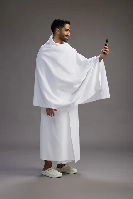 بورتريه لرجل سعودي بملابس الإحرام يبتسم ويلتقط صورة سيلفي, استخدام الهاتف المحمول, الحج والعمره في مكه, الجسم بالكامل, خلفيه رماديه