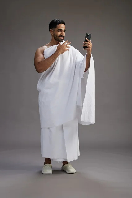 A portrait of a Saudi man in Ihram clothing smiling and taking a selfie, using a mobile phone, during Hajj and Umrah in Mecca, full body, gray background.