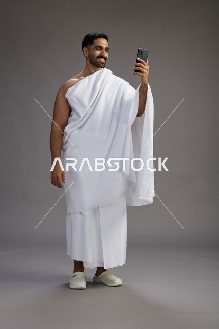 بورتريه لرجل سعودي بملابس الإحرام يبتسم ويلتقط صورة سيلفي, استخدام الهاتف المحمول, الحج والعمره في مكه, الجسم بالكامل, خلفيه رماديه