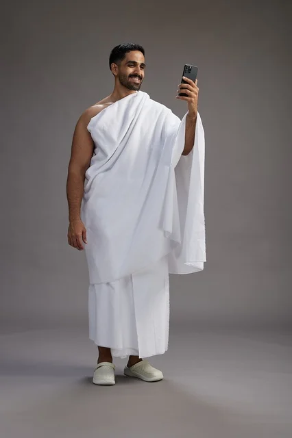 بورتريه لرجل سعودي بملابس الإحرام يبتسم ويلتقط صورة سيلفي, استخدام الهاتف المحمول, الحج والعمره في مكه, الجسم بالكامل, خلفيه رماديه