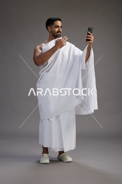بورتريه لرجل سعودي بملابس الإحرام يبتسم ويلتقط صورة سيلفي, استخدام الهاتف المحمول, الحج والعمره في مكه, الجسم بالكامل, خلفيه رماديه