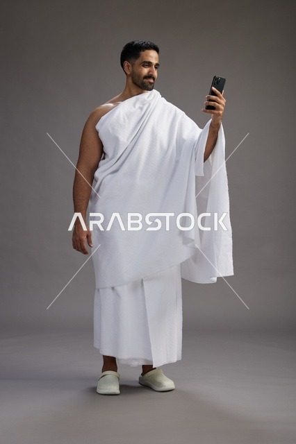 A portrait of a Saudi man in Ihram clothing smiling and taking a selfie, using a mobile phone, during Hajj and Umrah in Mecca, full body, gray background.