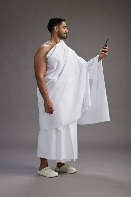 A portrait of a Saudi man in Ihram clothing smiling and taking a selfie, using a mobile phone, during Hajj and Umrah in Mecca, full body, gray background.
