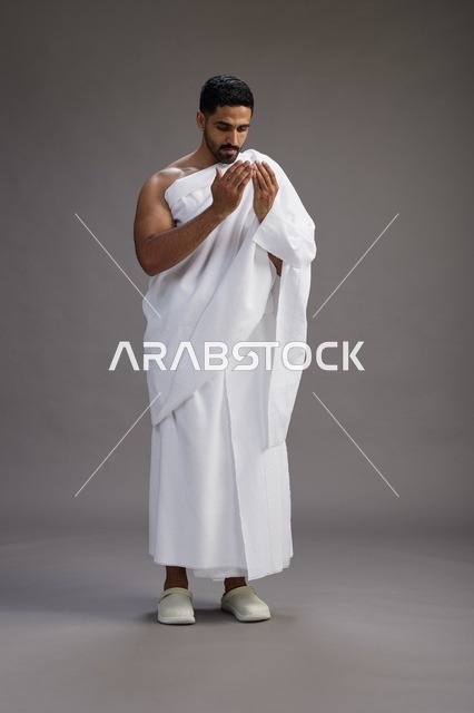 بورتريه لرجل سعودي بملابس الإحرام يدعو رافعاً يديه, الدعاء والتضرع لله , كامل الجسم, خلفيه رماديه