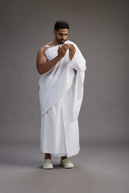 A portrait of a Saudi man in Ihram clothing, praying with his hands raised, supplicating and beseeching God, full body, gray background.