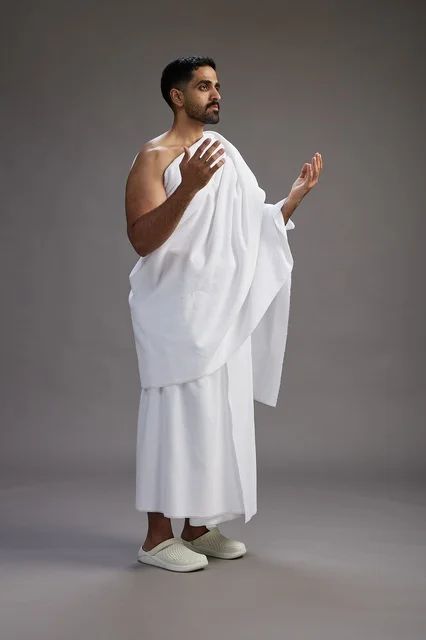 A portrait of a Saudi man in Ihram clothing, praying with his hands raised, supplicating and beseeching God, full body, gray background.