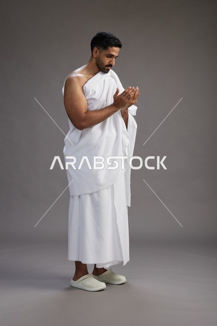 بورتريه لرجل سعودي بملابس الإحرام يدعو رافعاً يديه, الدعاء والتضرع لله , كامل الجسم, خلفيه رماديه