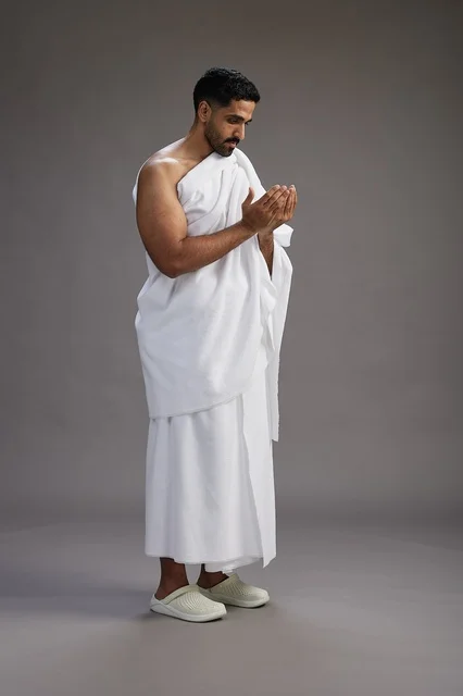 A portrait of a Saudi man in Ihram clothing, praying with his hands raised, supplicating and beseeching God, full body, gray background.