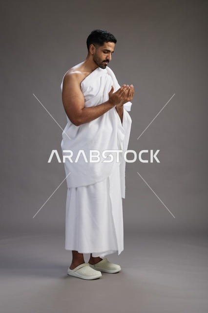 بورتريه لرجل سعودي بملابس الإحرام يدعو رافعاً يديه, الدعاء والتضرع لله , كامل الجسم, خلفيه رماديه
