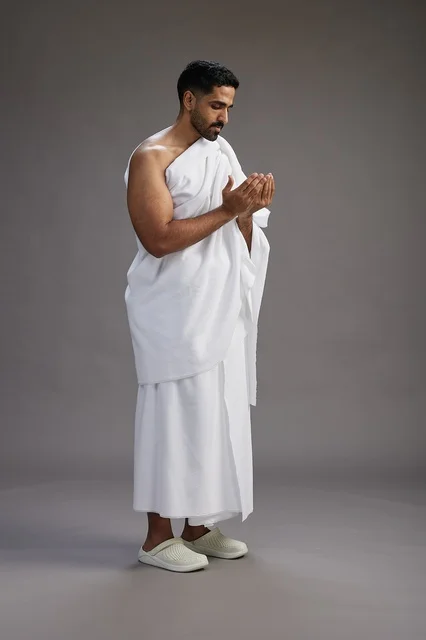A portrait of a Saudi man in Ihram clothing, praying with his hands raised, supplicating and beseeching God, full body, gray background.
