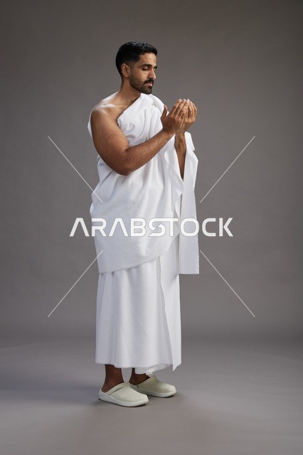بورتريه لرجل سعودي بملابس الإحرام يدعو رافعاً يديه, الدعاء والتضرع لله , كامل الجسم, خلفيه رماديه