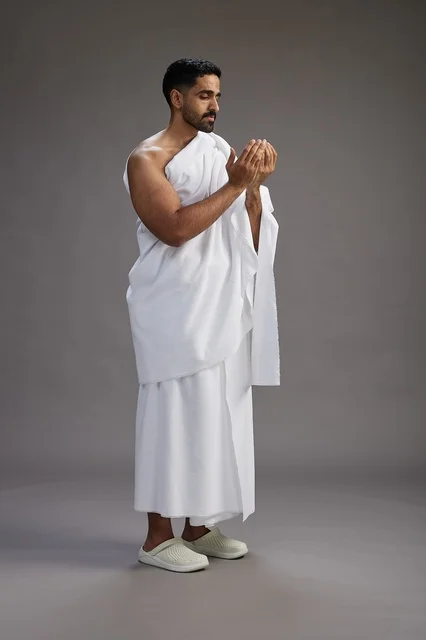 A portrait of a Saudi man in Ihram clothing, praying with his hands raised, supplicating and beseeching God, full body, gray background.