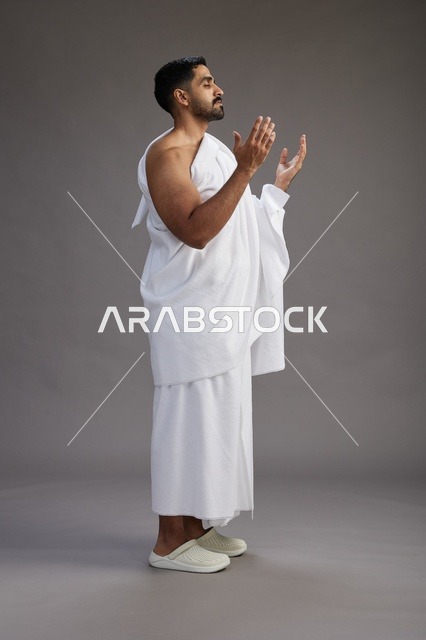 بورتريه لرجل سعودي بملابس الإحرام يدعو رافعاً يديه, الدعاء والتضرع لله , كامل الجسم, خلفيه رماديه