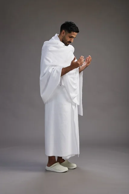 A portrait of a Saudi man in Ihram clothing, praying with his hands raised, supplicating and beseeching God, full body, gray background.