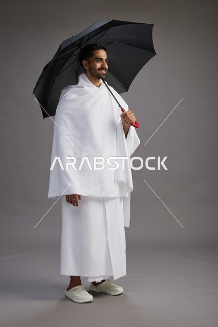 بورتريه لحاج  عربي سعودي  خليجي بملابس الإحرام يحمل مظله, الذهاب لاداء مناسك الحج والعمره,  كامل الجسم, خلفيه رماديه