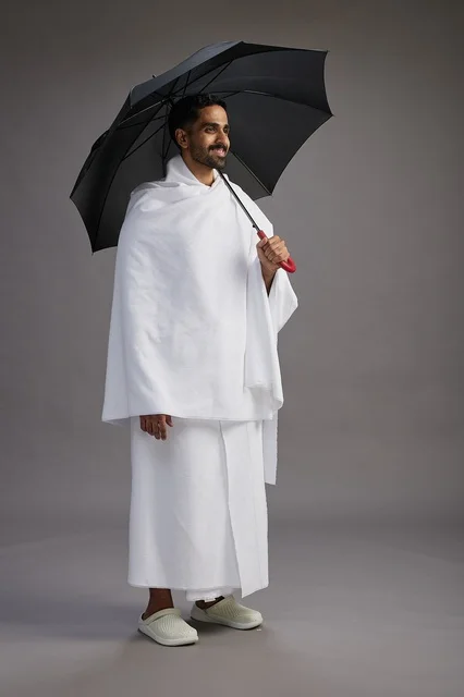A portrait of an Arab Saudi Gulf pilgrim in Ihram clothing holding an umbrella, going to perform the rituals of Hajj and Umrah, full body, gray background.