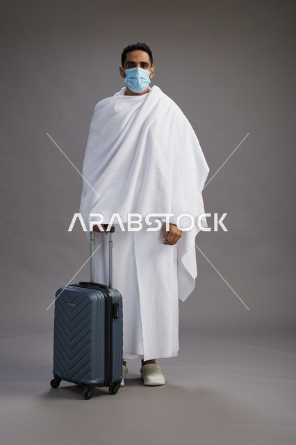 رجل عربي بملابس الإحرام يحمل حقيبة سفر وكمامة طبية