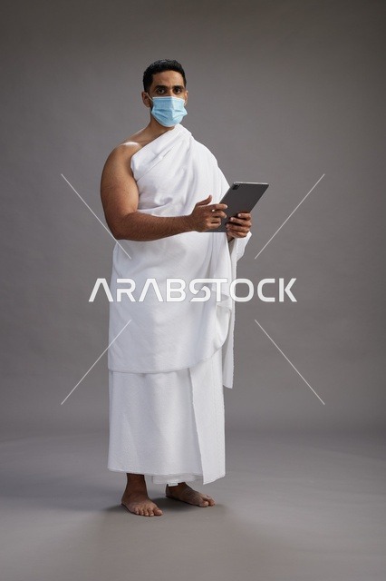 رجل سعودي بملابس الإحرام يحمل جهاز لوحي مع كمامة طبية