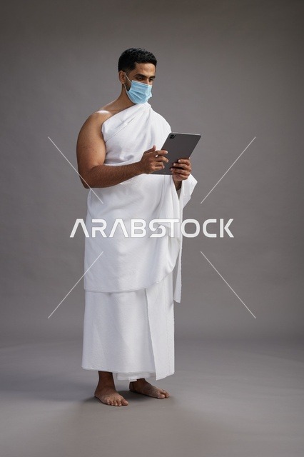 رجل سعودي بملابس الإحرام يستخدم جهاز لوحي بالكمامة