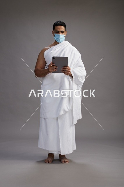 رجل عربي بملابس الإحرام مع كمامة وجهاز لوحي