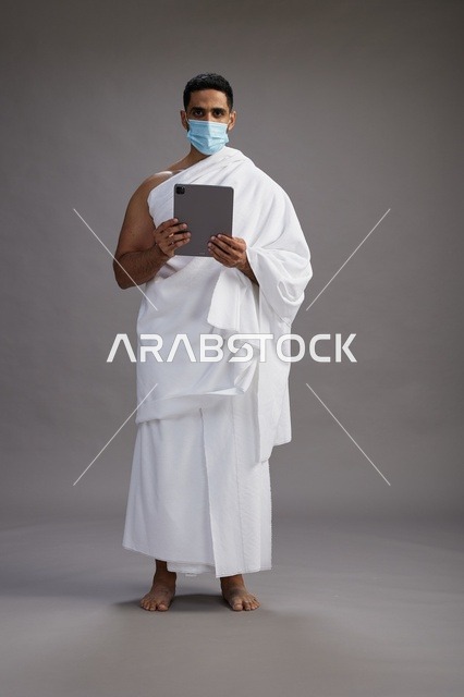 رجل سعودي بملابس الإحرام يرتدي كمامة ويحمل تابلت