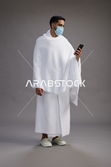 حاج سعودي بملابس الإحرام يستخدم الجوال مع الكمامة