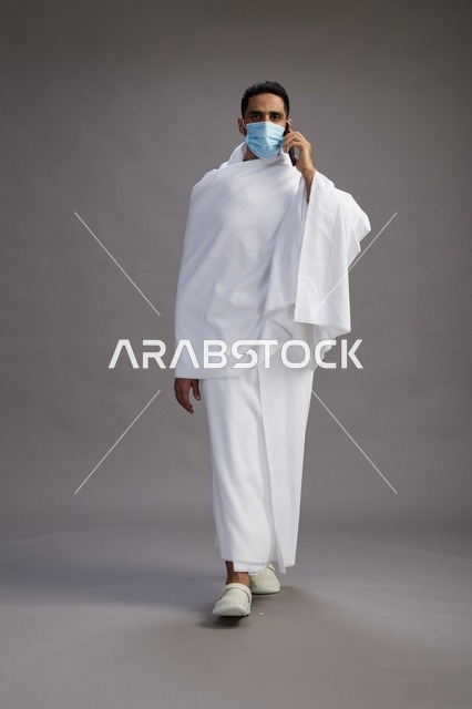 رجل سعودي بملابس الإحرام والكمامة يستخدم الهاتف الذكي