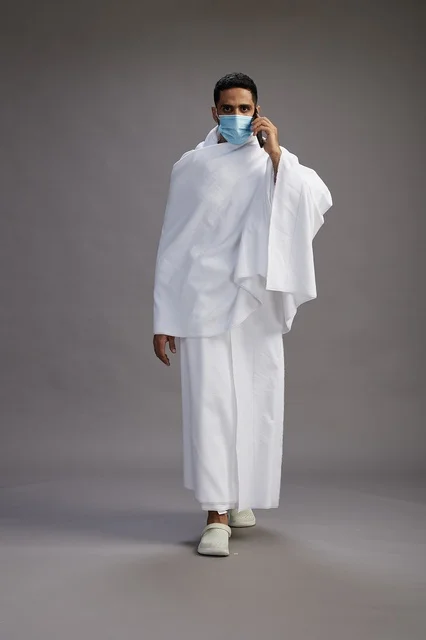 رجل عربي بملابس الإحرام يرتدي كمامة ويستخدم الجوال