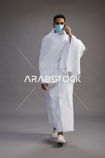 رجل سعودي بملابس الإحرام يرتدي كمامة مع هاتف ذكي