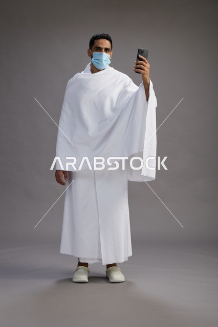 حاج سعودي بملابس الإحرام يستخدم الجوال مع الكمامة