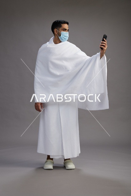 حاج سعودي بملابس الإحرام يستخدم الجوال مع كمامة