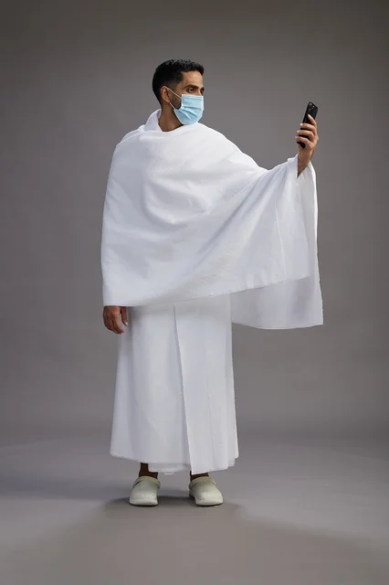 حاج سعودي بملابس الإحرام يستخدم الجوال مع كمامة