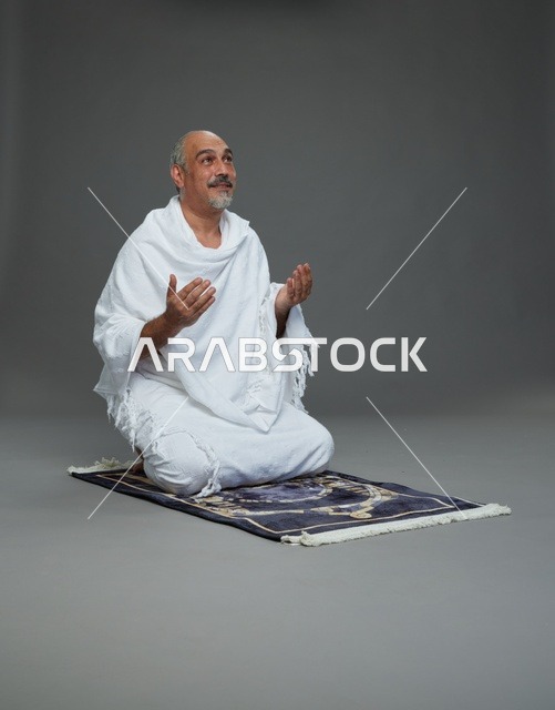 بورتريه لحاج كبير السن عربي سعودي خليجي بملابس الإحرام  يجلس على سجادة صلاه ويرفع يديه بالدعاء, اداء مناسك الحج والعمره , خلفيه رماديه