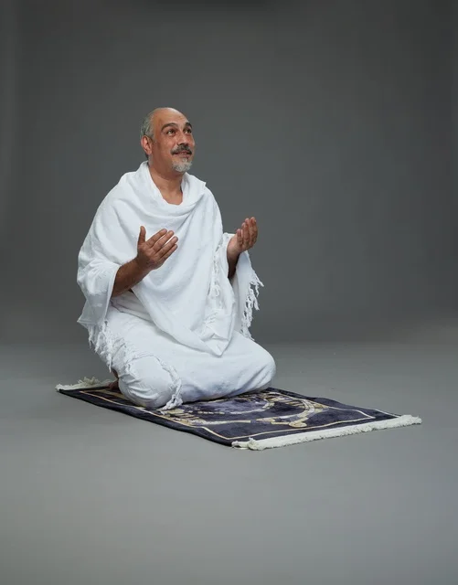بورتريه لحاج كبير السن عربي سعودي خليجي بملابس الإحرام  يجلس على سجادة صلاه ويرفع يديه بالدعاء, اداء مناسك الحج والعمره , خلفيه رماديه