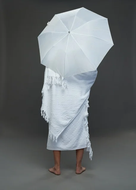 بورتريه لرجل سعودي بملابس الإحرام يحمل مظلة بيضاء أثناء أداء المناسك, ايماءات واشارات مختلفه, كامل الجسم, خلفيه رماديه