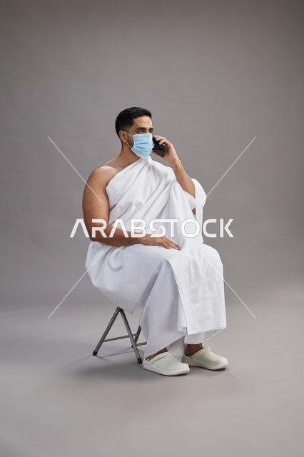 رجل سعودي بملابس الإحرام يجلس مرتدياً كمامة أثناء مكالمة هاتفية