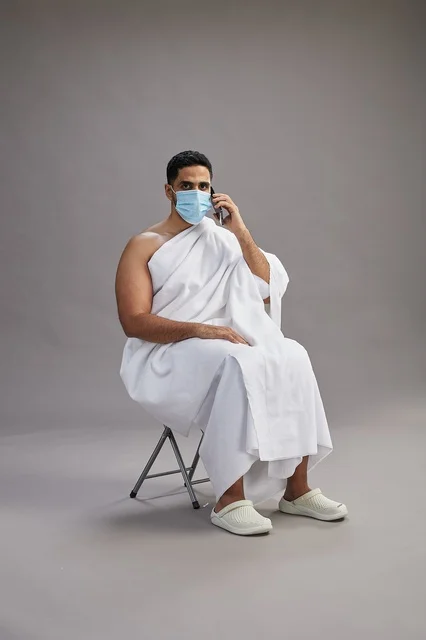 رجل سعودي بملابس الإحرام يجلس مرتدياً كمامة أثناء مكالمة هاتفية