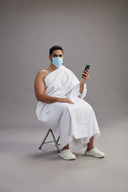رجل سعودي بملابس الإحرام يجلس مرتدياً كمامة ويستخدم الهاتف المحمول