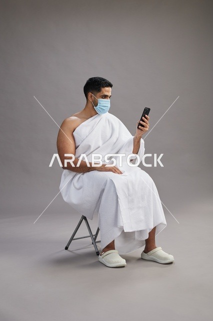 رجل سعودي بملابس الإحرام يجلس مرتدياً كمامة ويستخدم الهاتف المحمول
