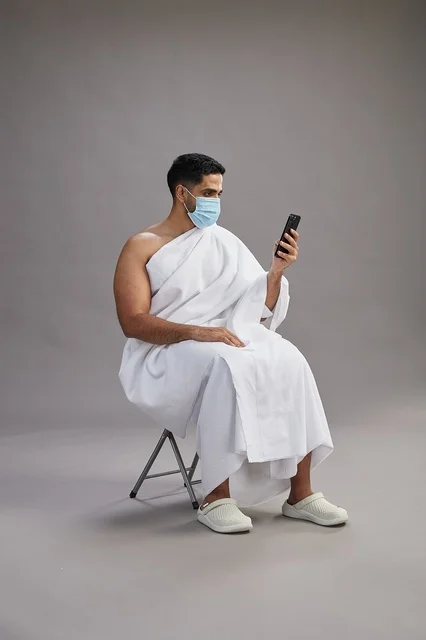 رجل سعودي بملابس الإحرام يجلس مرتدياً كمامة ويستخدم الهاتف المحمول