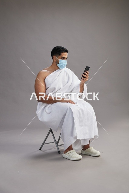 رجل سعودي بملابس الإحرام يجلس مرتدياً كمامة ويستخدم الهاتف المحمول