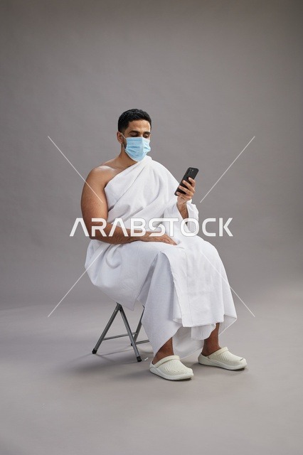 رجل سعودي بملابس الإحرام يجلس مرتدياً كمامة ويستخدم الهاتف المحمول