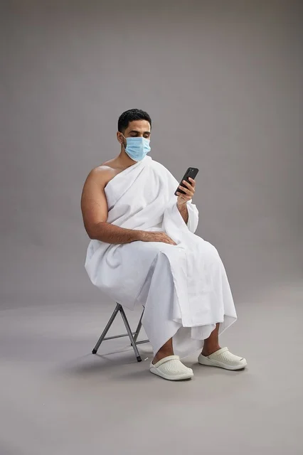 Saudi man in ihram sitting and using smartphone with face mask