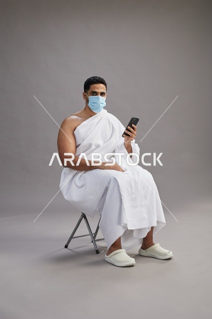 رجل سعودي بملابس الإحرام يجلس مرتدياً كمامة ويستخدم الهاتف المحمول