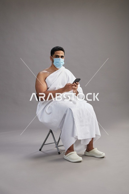 رجل سعودي بملابس الإحرام يجلس مرتدياً كمامة ويستخدم الهاتف المحمول
