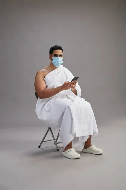 Saudi man in ihram sitting and using smartphone with face mask