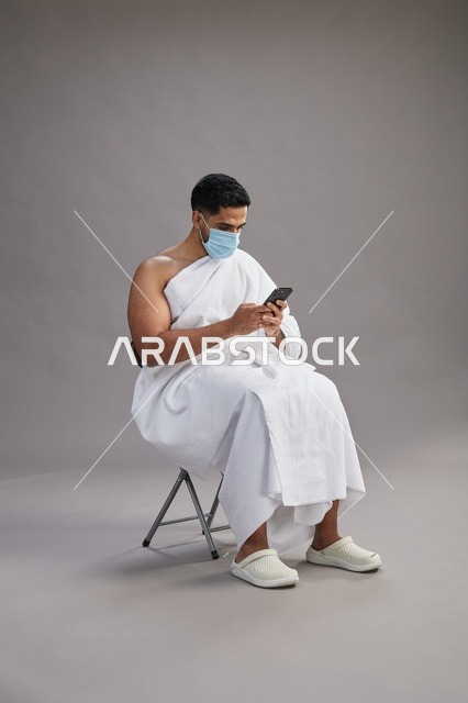 Saudi man in ihram sitting and using smartphone with face mask