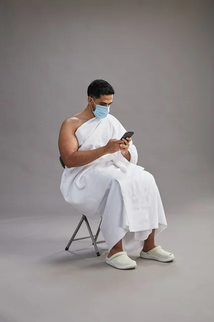 Saudi man in ihram sitting and using smartphone with face mask