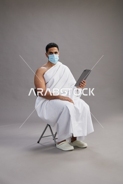 رجل سعودي بملابس الإحرام يستخدم جهاز لوحي وكمامة